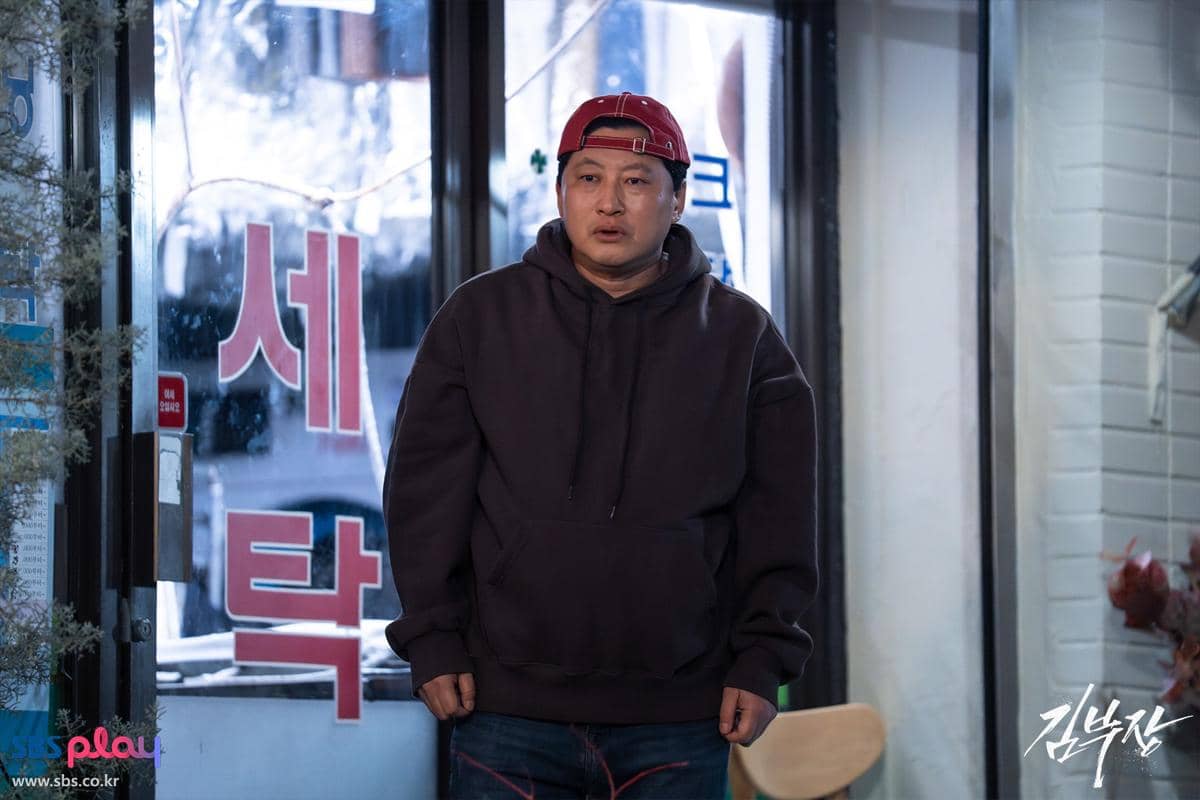 Seorang pria mengenakan hoodie gelap dan topi merah terbalik berdiri di depan pintu kaca dengan tulisan Korea di latar belakang.
