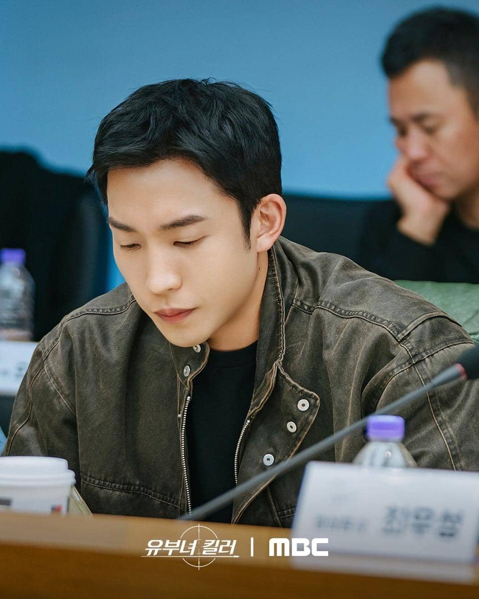 Seorang aktor mengenakan jaket cokelat duduk di meja membaca naskah saat sesi pembacaan untuk drama A Bona Fide Killer produksi MBC.