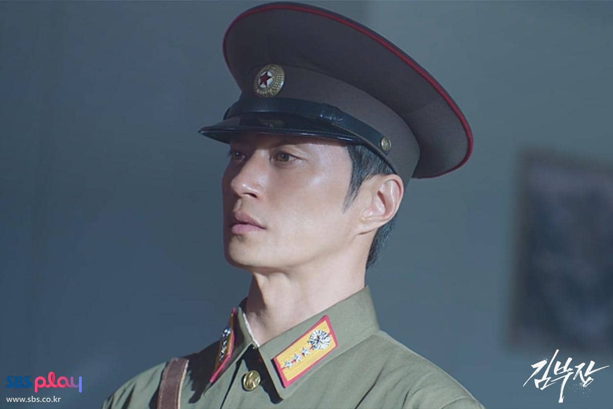 Seorang pria mengenakan seragam militer berwarna hijau dengan topi pet dan lambang bintang, dalam adegan drama Agent Kim Reactivated.