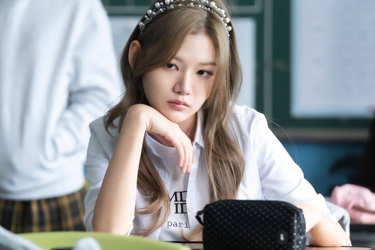 Seorang siswi berambut panjang mengenakan bando mutiara duduk di kelas dengan ekspresi serius dalam adegan drama Agent Kim Reactivated.