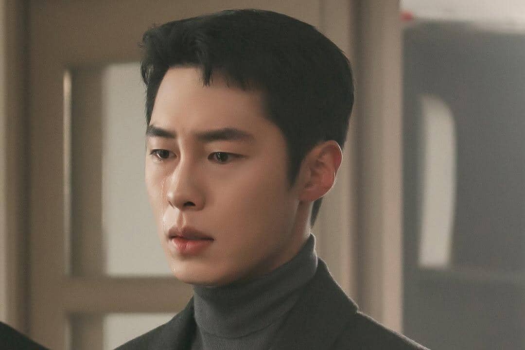 Seorang pria berwajah serius mengenakan turtleneck abu-abu dan jas gelap dalam adegan drama Doctor on the Edge.