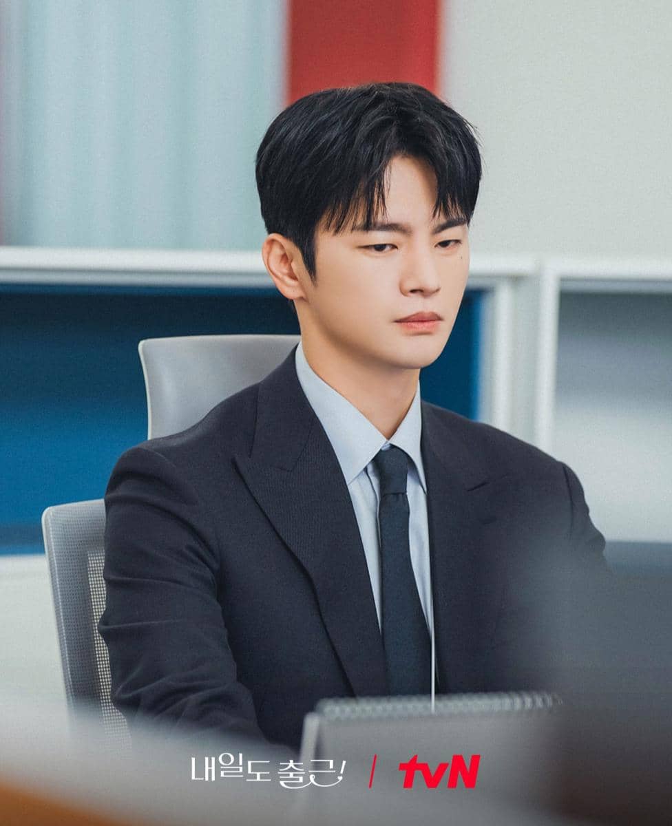 Seorang pria mengenakan setelan jas dan dasi duduk di kursi kantor dengan ekspresi serius di cuplikan drama See You At Work Tomorrow.