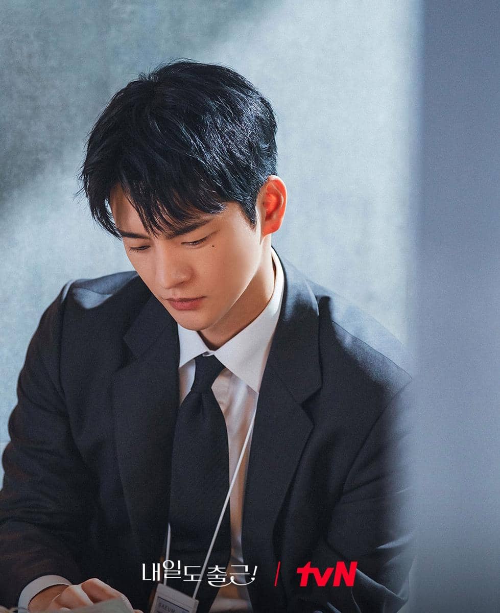 Seorang pria mengenakan setelan jas hitam dan dasi sedang duduk dengan ekspresi serius dalam cuplikan drama See You At Work Tomorrow.