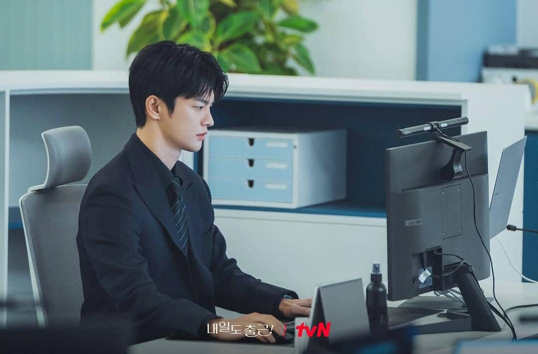 Seorang pria mengenakan setelan jas hitam sedang bekerja di depan komputer di ruang kantor modern dengan suasana tenang.