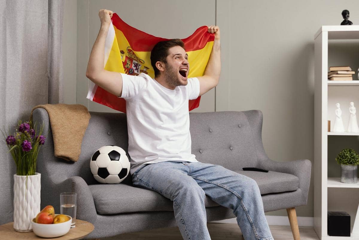 Pria duduk di sofa sambil mengibarkan bendera Spanyol dan bersorak gembira menonton pertandingan sepak bola di ruang tamu.
