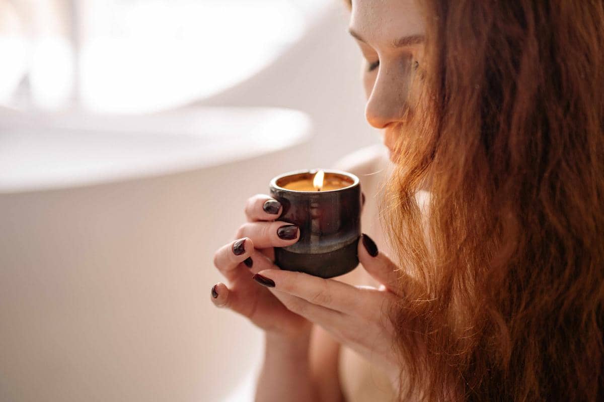Seorang perempuan dengan rambut panjang memegang lilin aromaterapi yang menyala sambil menikmati aromanya di ruangan terang.