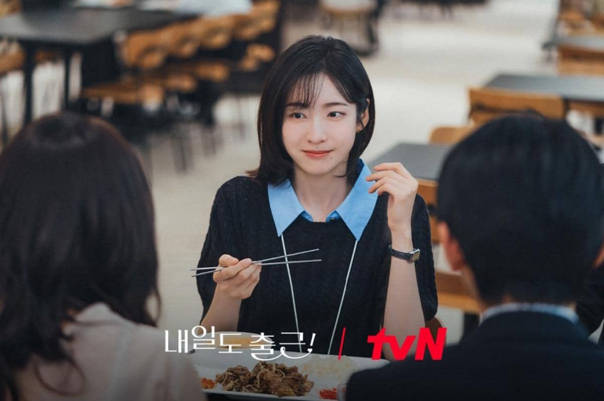 Seorang wanita muda tersenyum sambil memegang sumpit di meja makan bersama dua orang lainnya dalam adegan drama See You at Work Tomorrow.