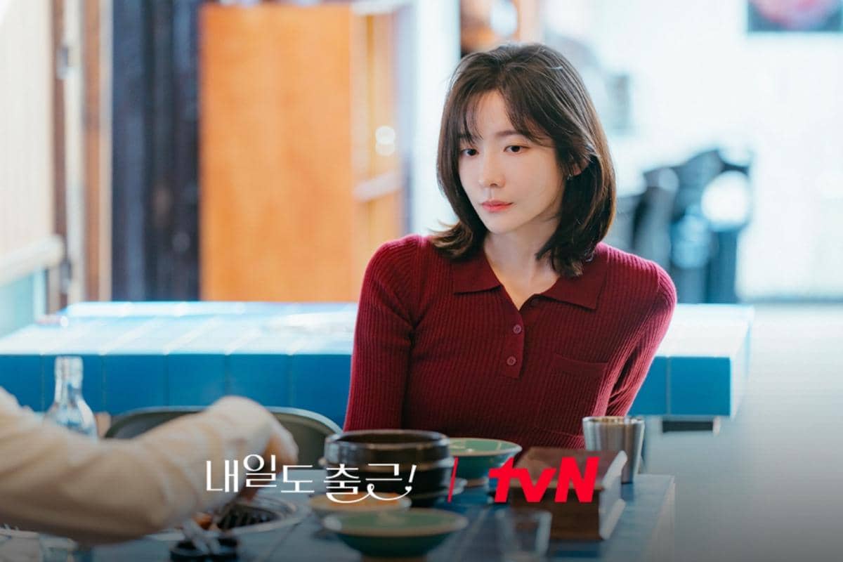 Seorang wanita berambut pendek mengenakan sweater merah duduk di meja restoran dengan ekspresi serius dalam adegan drama See You at Work Tomorrow.