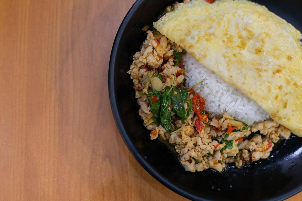 Semangkuk nasi ayam cincang pedas gurih disajikan dengan telur dadar di atas piring hitam di atas meja kayu.