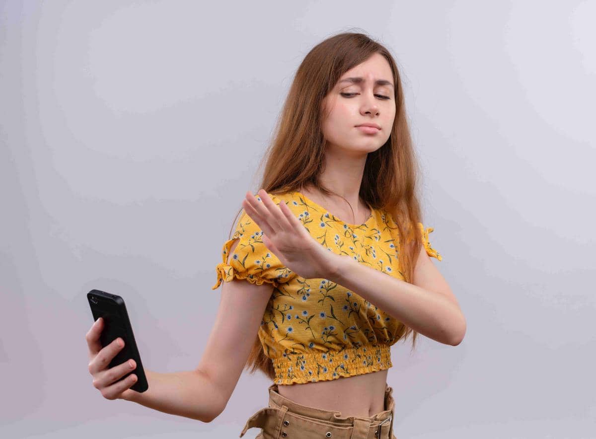 Seorang perempuan mengenakan blus kuning bermotif bunga memegang ponsel sambil mengangkat tangan seolah menolak untuk melihat layar.