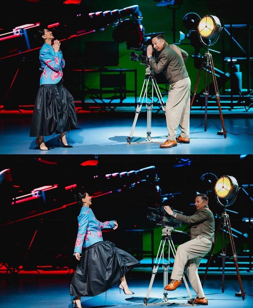 Seorang pria mengoperasikan kamera film di atas tripod sementara seorang wanita bergaya ceria di depan panggung dengan pencahayaan teater berwarna.
