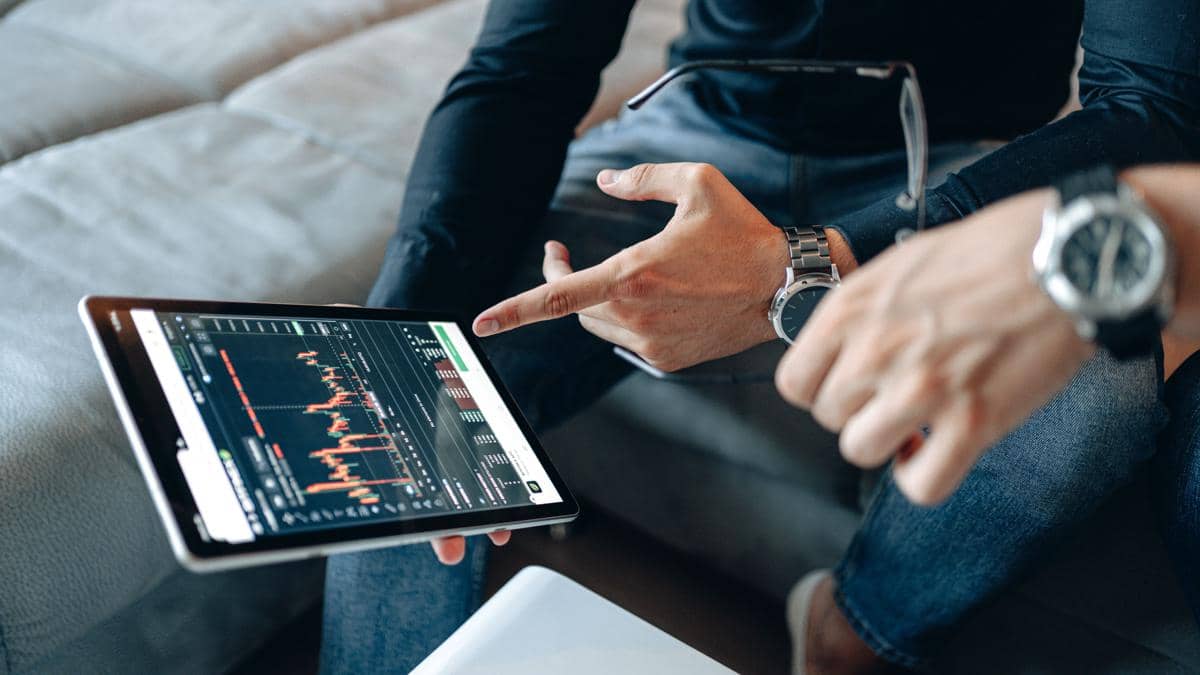 Dua orang sedang menganalisis grafik perdagangan saham di layar tablet dengan tampilan grafik harga dan data pasar.