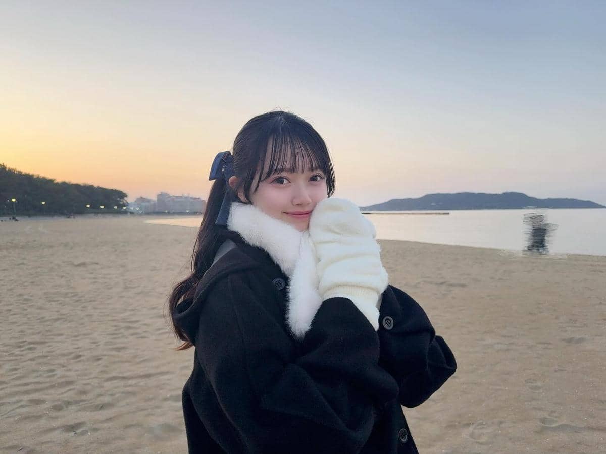 Mei Hanada berjaket hitam dan sarung tangan putih tersenyum di pantai saat matahari terbenam dengan latar laut dan bukit.