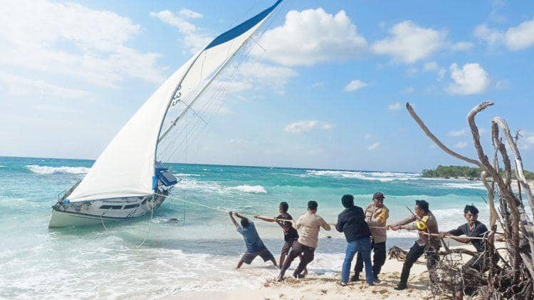 Beberapa orang menarik tali untuk membantu kapal pesiar berbendera Australia yang terdampar di Pantai Duile, Rote Ndao, dengan ombak dan langit cerah di latar belakang.