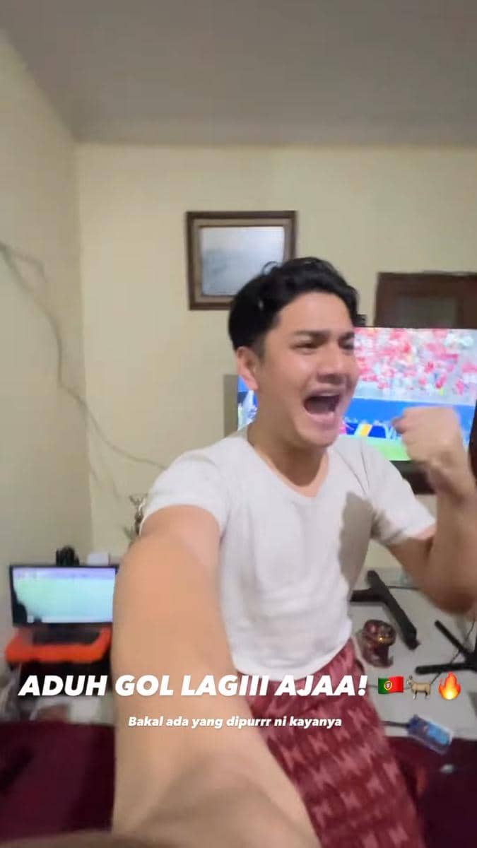 Seorang pria mengenakan kaus putih dan sarung merah berteriak gembira di depan televisi yang menayangkan pertandingan sepak bola.