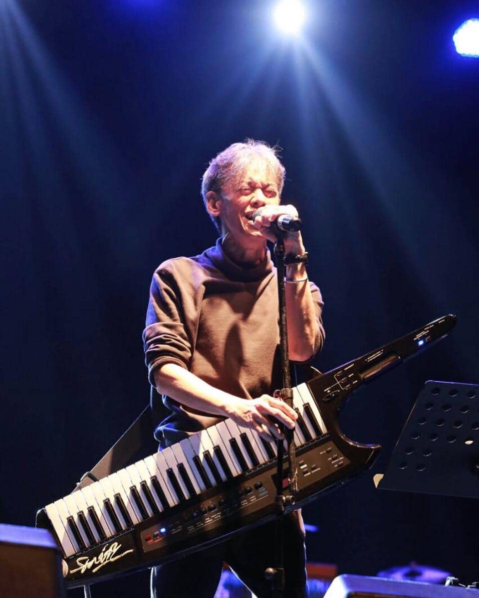 Seorang musisi sedang bernyanyi sambil memainkan keytar di atas panggung dengan pencahayaan sorot biru dan putih yang dramatis.