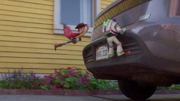 Woody dan Buzz Lightyear berpegangan di bagian belakang mobil yang sedang melaju di depan rumah berwarna kuning dengan taman bunga.