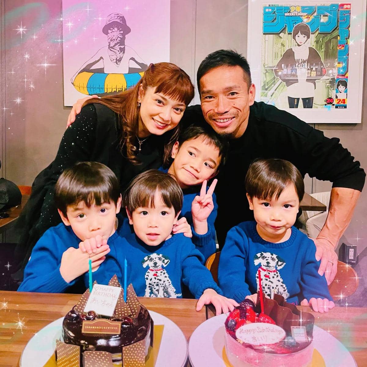 Sebuah keluarga dengan tiga anak kecil berpose bersama di depan dua kue ulang tahun di atas meja, semuanya tersenyum hangat.