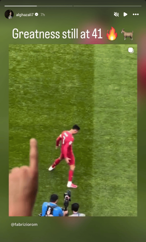 Seorang pemain sepak bola mengenakan seragam merah di lapangan hijau dengan penonton dan fotografer di tepi lapangan.