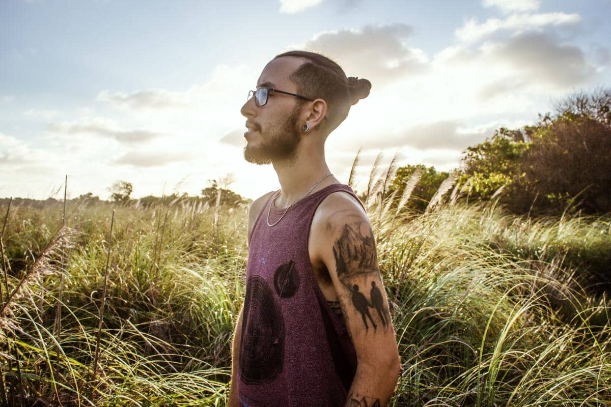 Pria berambut man bun mengenakan kacamata dan kaus tanpa lengan berdiri di tengah padang rumput saat matahari terbenam.