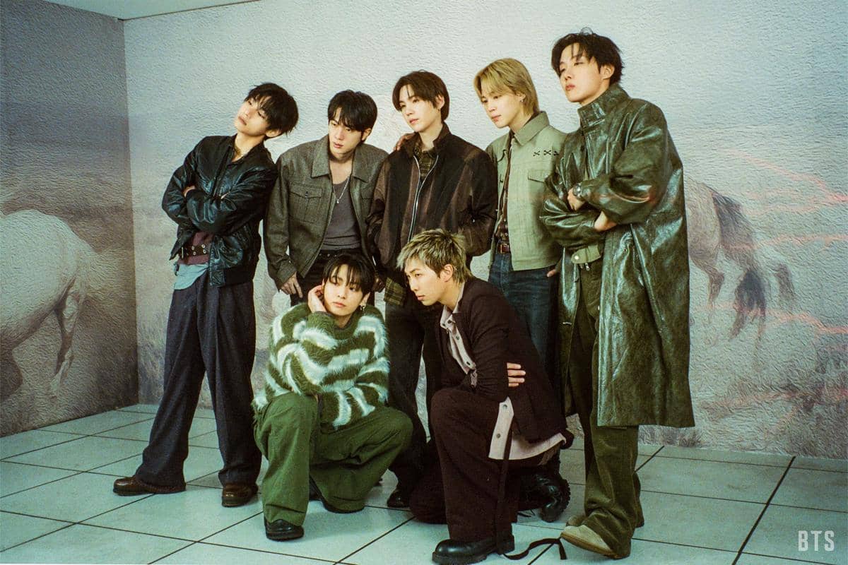 Tujuh anggota BTS berpose bersama dengan busana kasual dan jaket kulit dalam set video musik Merry Go Round berlatar dinding bergambar kuda.