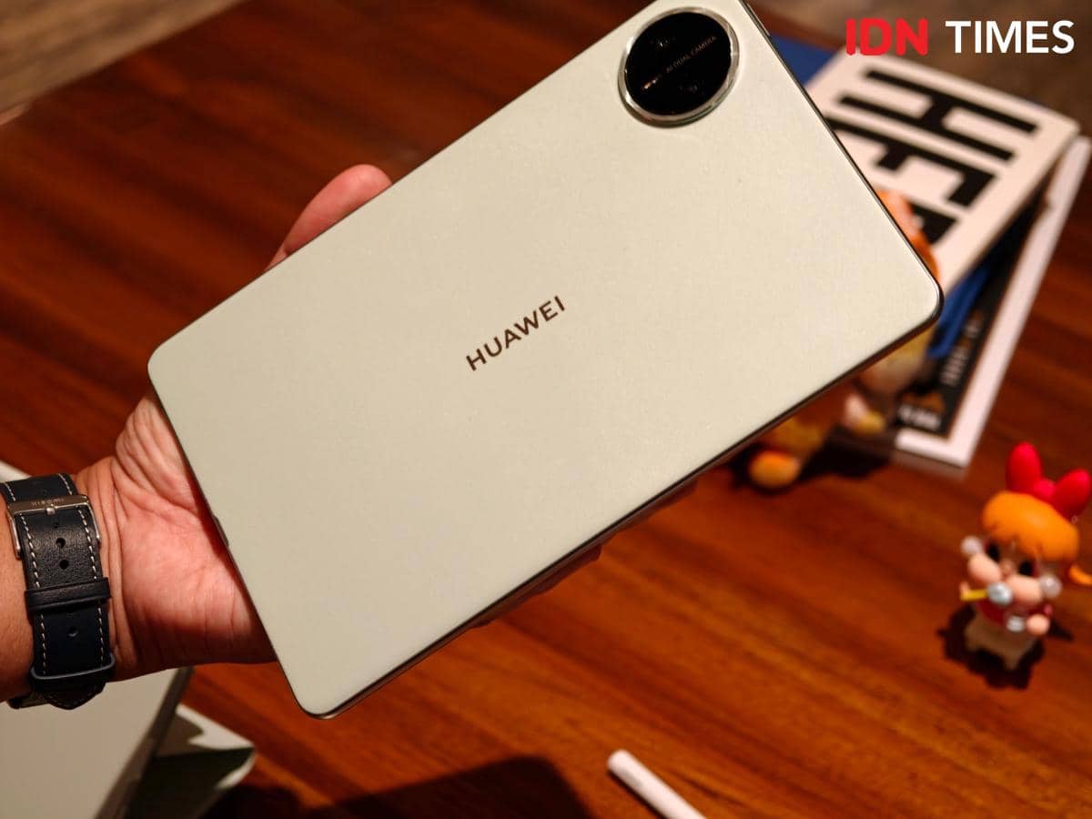 Seseorang memegang tablet HUAWEI MatePad Mini berwarna abu-abu muda dengan kamera belakang besar di atas meja kayu.