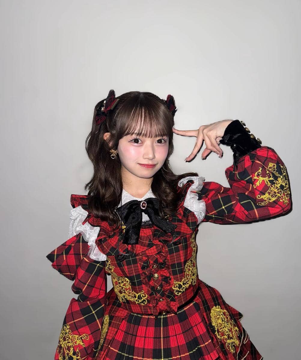 Mei Hanada berpose dengan tangan di dekat wajah, mengenakan gaun bergaya lolita bermotif kotak merah hitam dengan hiasan renda dan pita.