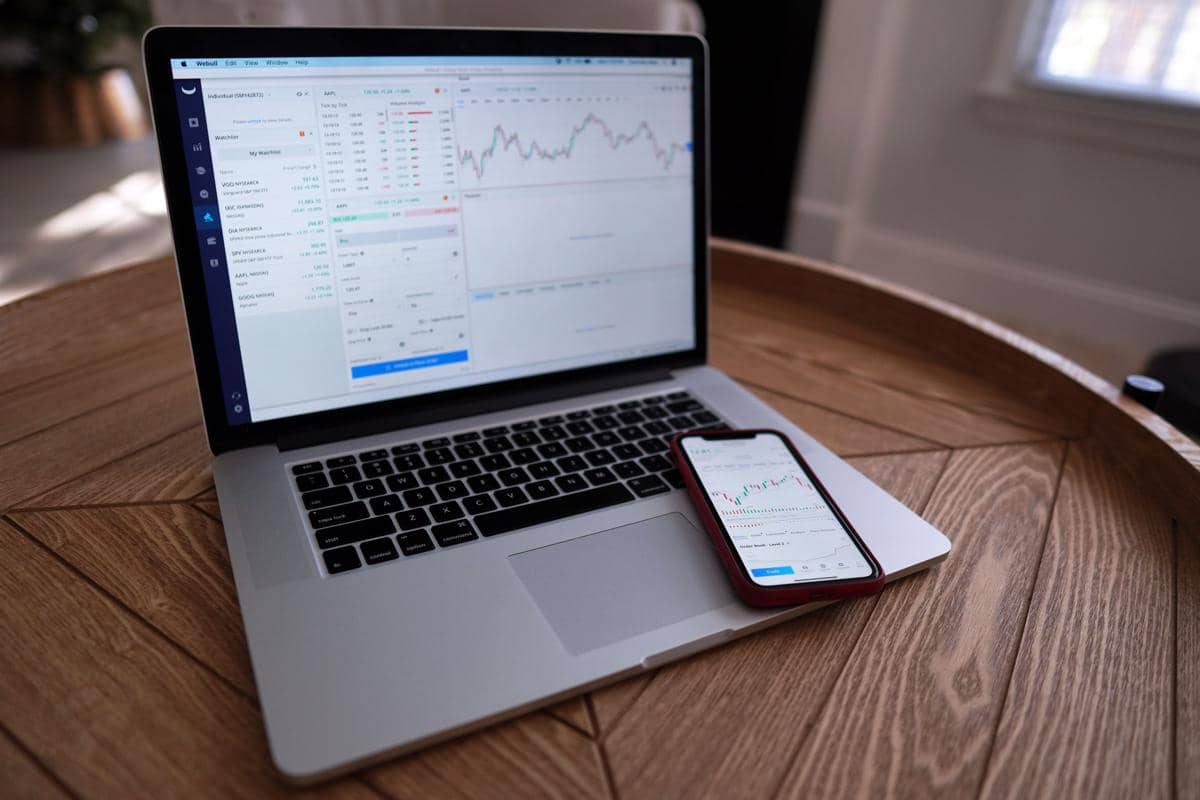 Laptop dan ponsel menampilkan grafik saham serta data perdagangan di atas meja kayu, menggambarkan aktivitas analisis pasar keuangan.