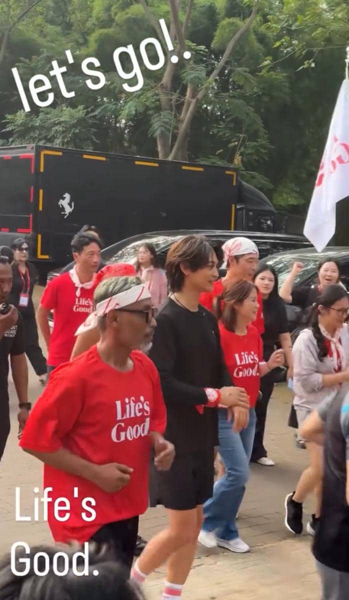 Seorang pria berlari bersama peserta lain mengenakan kaus merah bertuliskan 'Life's Good' di area luar ruangan dengan pepohonan rindang.