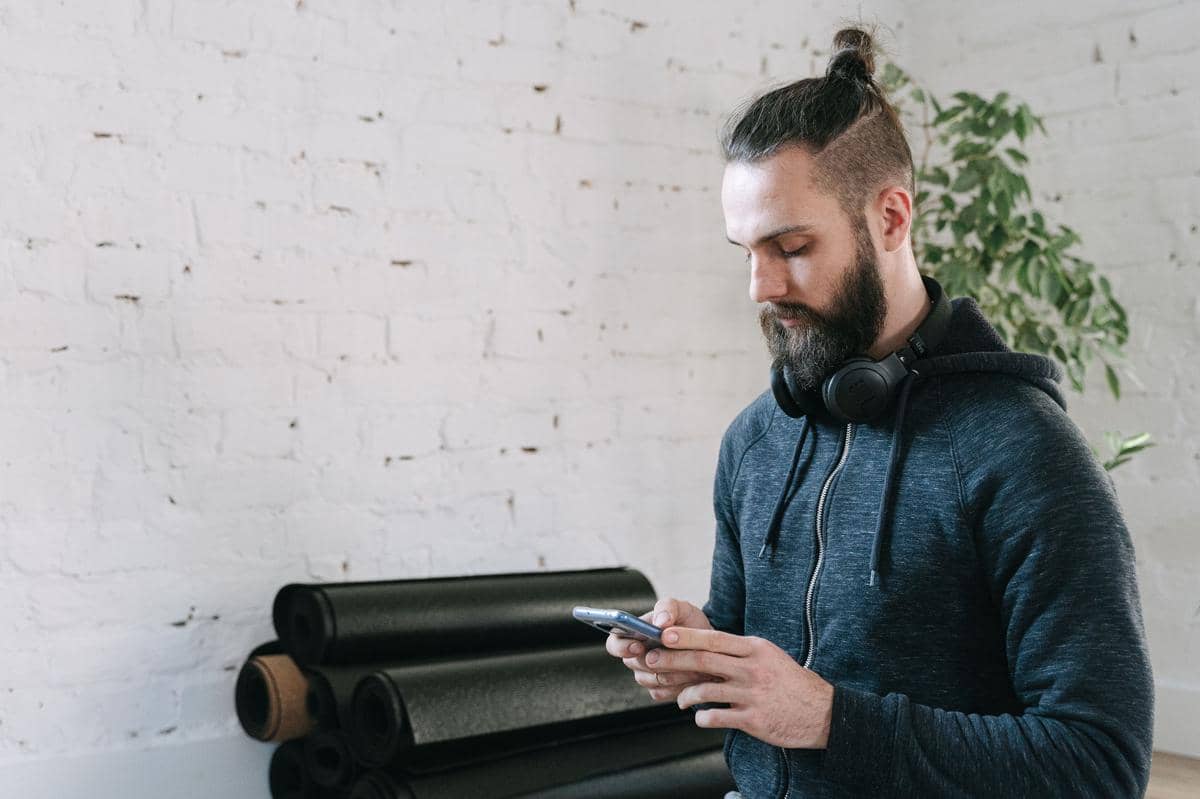 Pria berambut man bun dan berjanggut mengenakan hoodie biru tua serta headphone di lehernya sedang menggunakan ponsel.