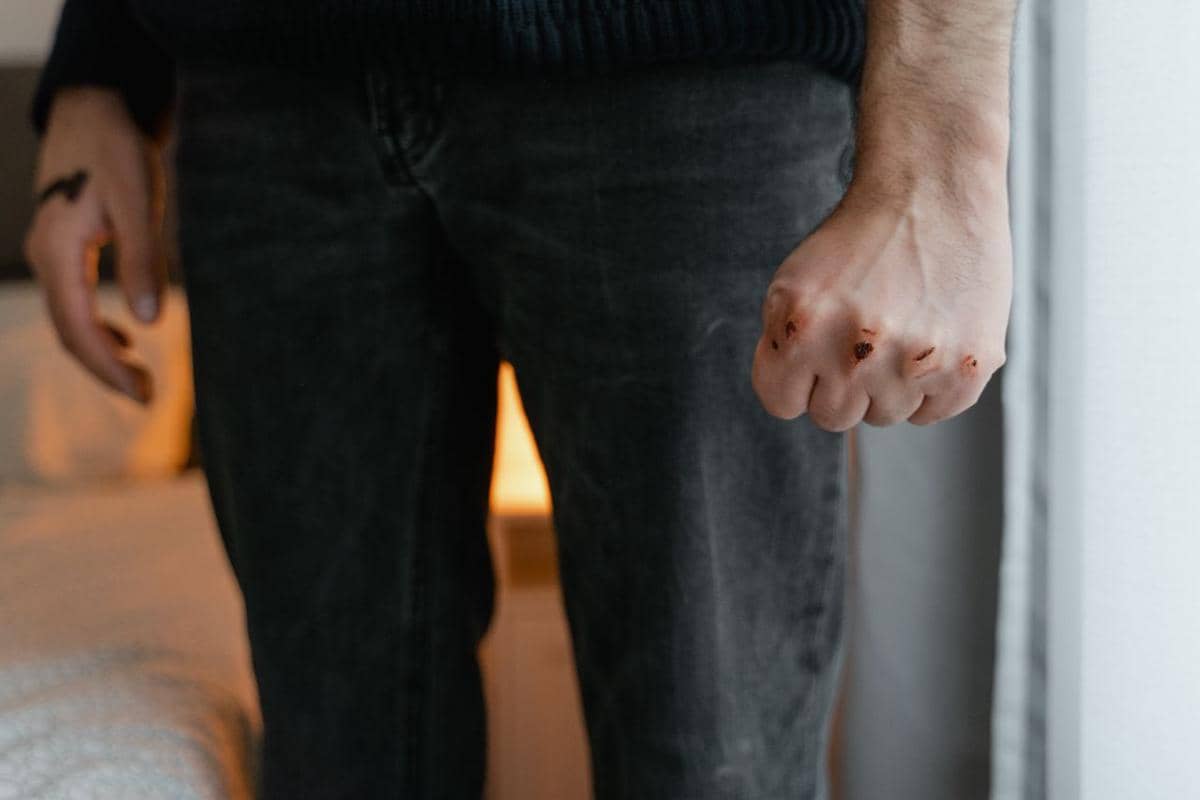 Tangan seseorang mengepal dengan luka di buku jari, mengenakan celana jeans gelap dan berdiri di dalam ruangan.