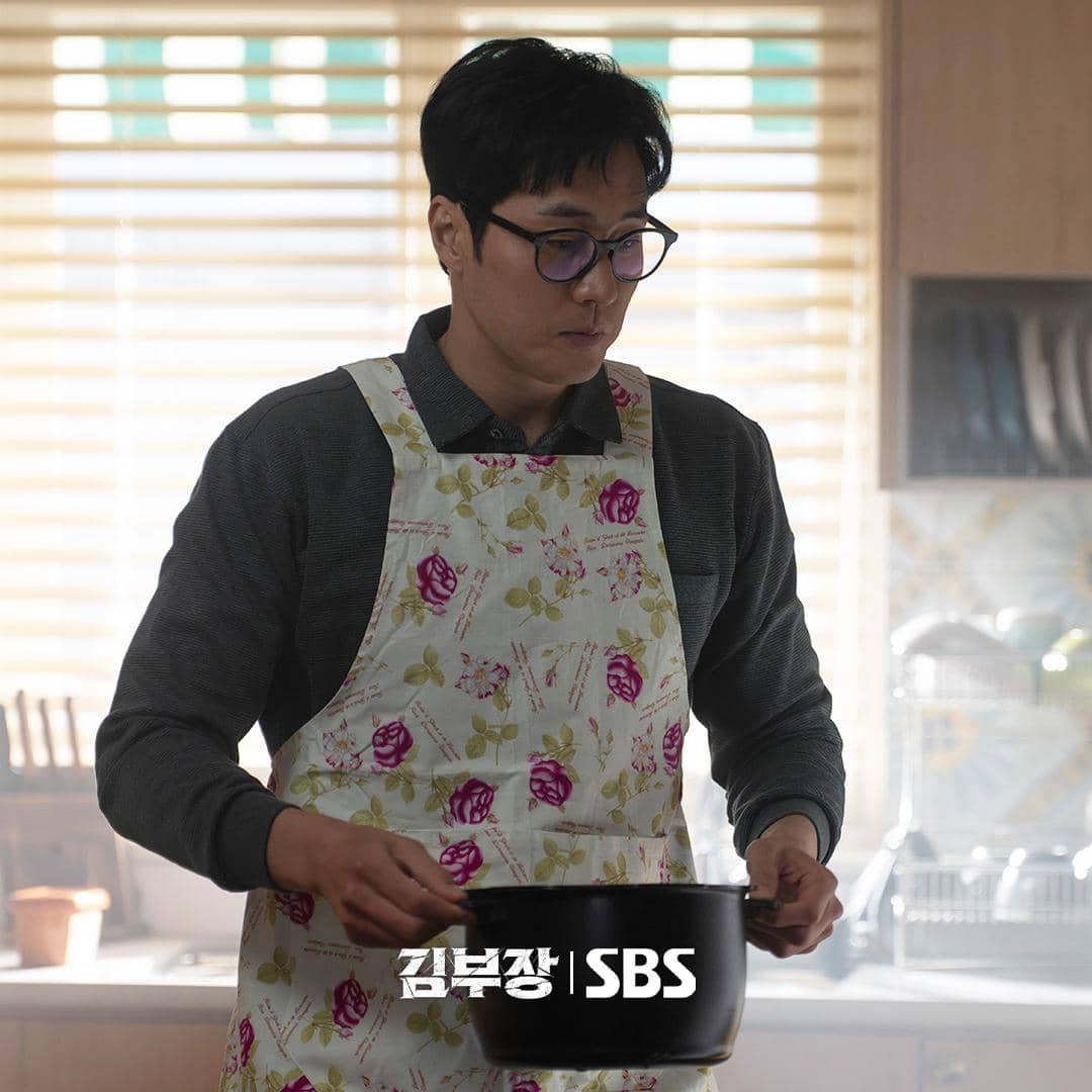 Seorang pria mengenakan celemek bermotif bunga sedang memegang panci di dapur dengan pencahayaan lembut dari jendela berkerai.