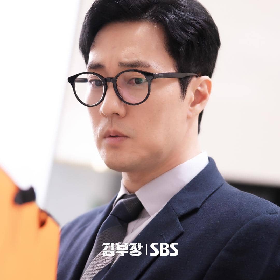 Seorang pria mengenakan setelan jas dan kacamata hitam bundar tampak serius dalam cuplikan drama Agent Kim Reactivated.