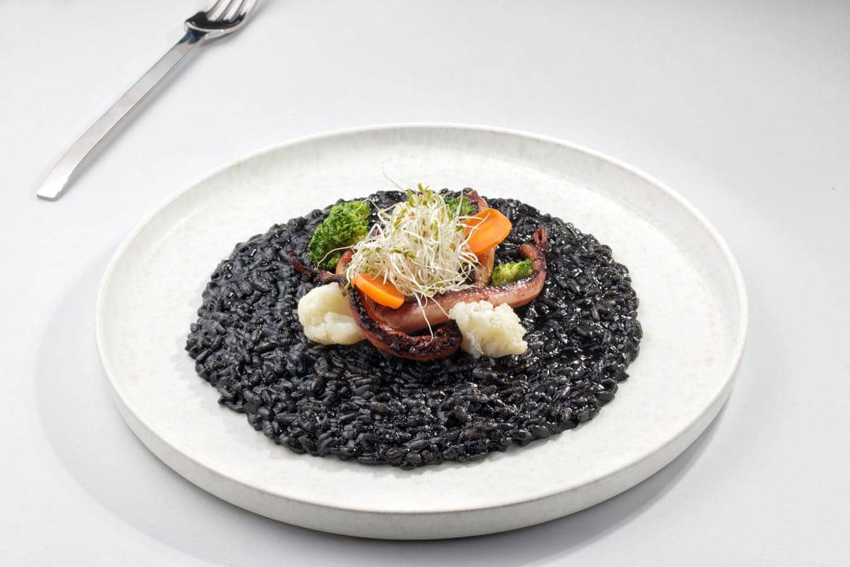 Risotto al nero di seppia berwarna hitam disajikan di piring putih dengan topping sayuran, gurita panggang, dan kecambah segar.