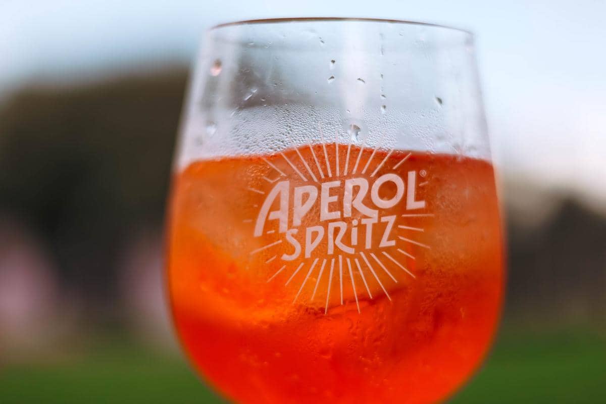 Segelas koktail Aperol Spritz berwarna oranye dengan es batu dan embun di permukaan gelas di latar luar ruangan.