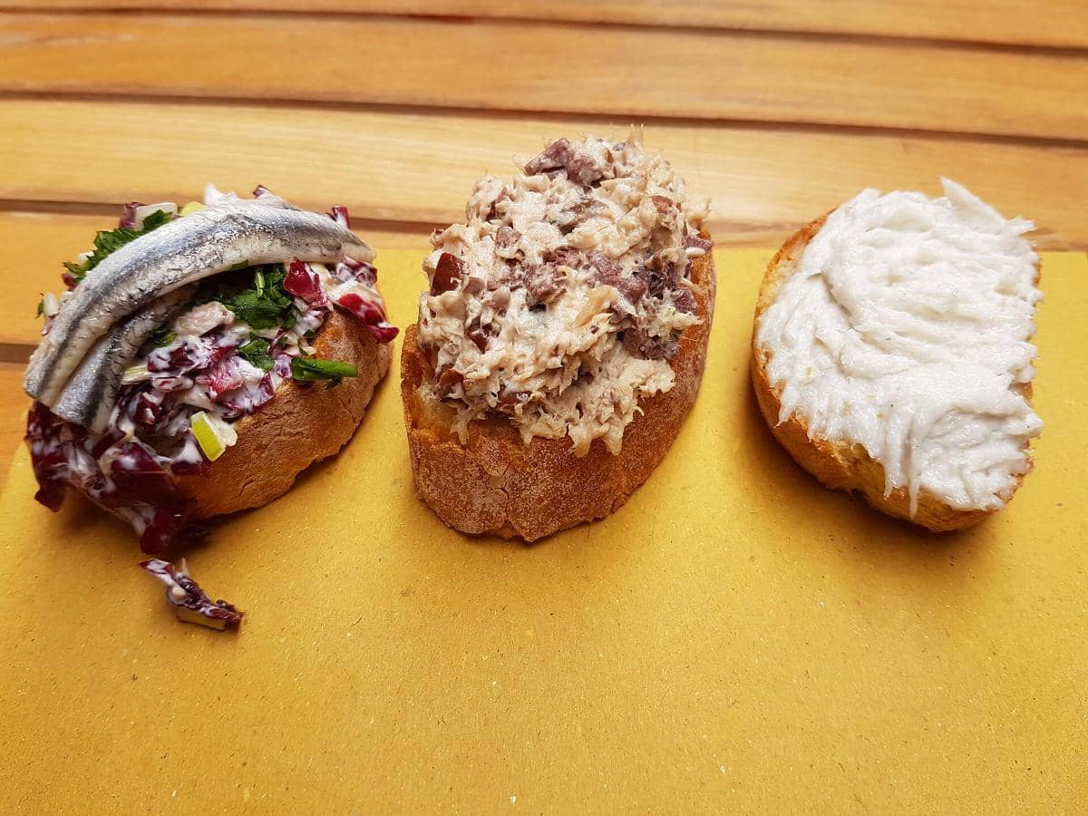 Tiga potong cicchetti khas Italia di atas meja kayu, masing-masing dengan topping ikan, krim, dan campuran sayuran segar.