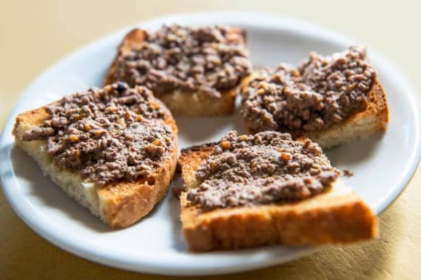 Empat potong crostini dengan olesan pate cokelat disajikan di atas piring putih di atas meja kayu berwarna terang.