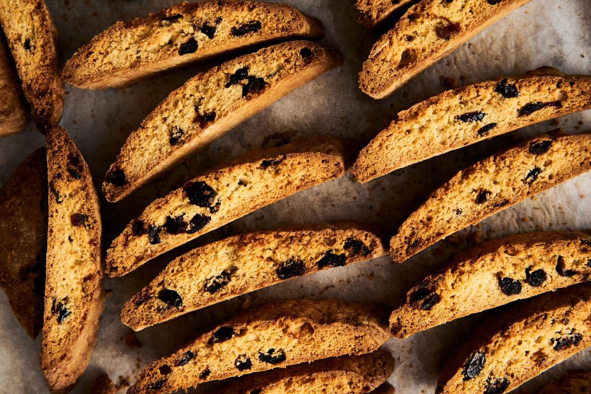 Beberapa potong biscotti berwarna keemasan dengan potongan buah kering tersusun di atas permukaan kertas panggang.