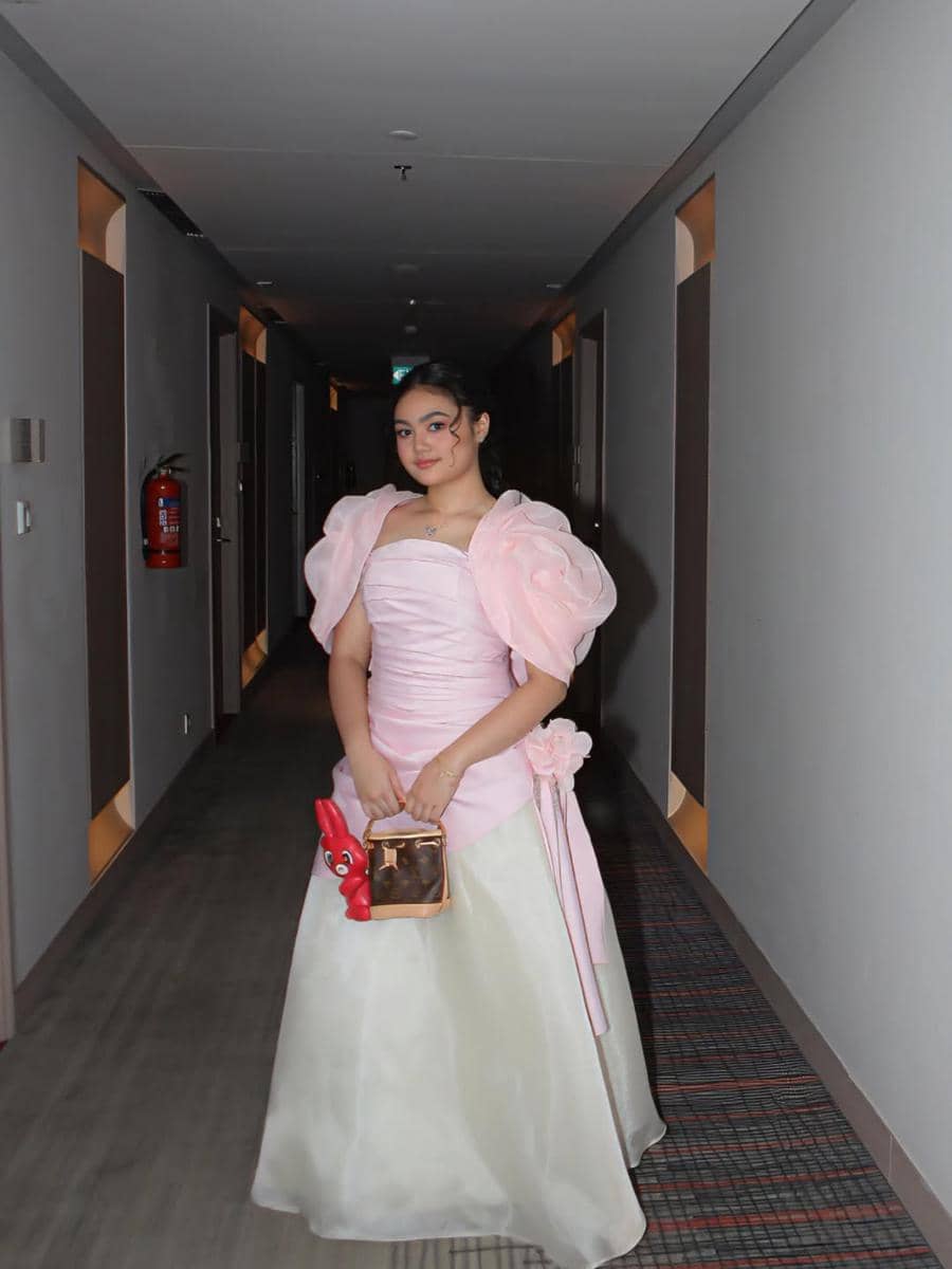 Seorang remaja mengenakan gaun prom berwarna pink dan putih dengan tas kecil di tangan, berdiri di lorong gedung yang diterangi lampu.