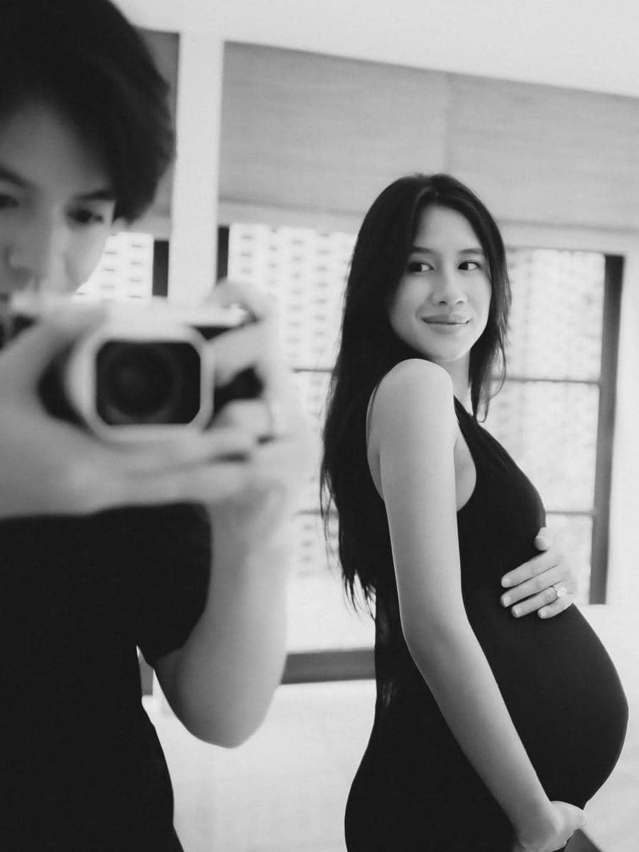 Seorang wanita hamil mengenakan gaun hitam berpose sambil tersenyum di depan kamera dalam pemotretan maternity hitam putih.