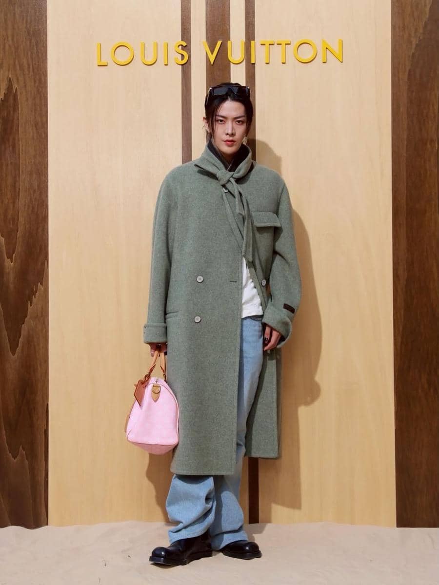 Seorang model berpose di depan latar bertuliskan Louis Vuitton mengenakan mantel hijau, celana longgar, dan tas tangan pink.