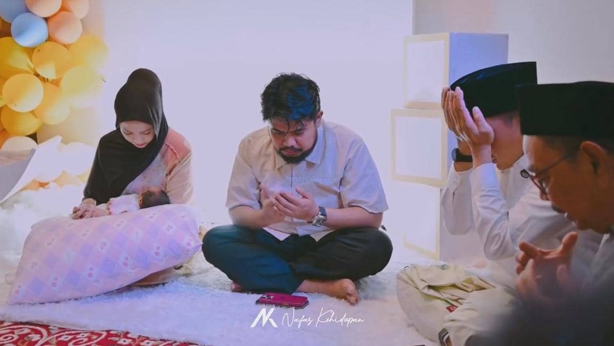 Suasana acara akikah dengan seorang ibu menggendong bayi, ayah dan tamu laki-laki berdoa bersama di ruangan berhias balon warna pastel.