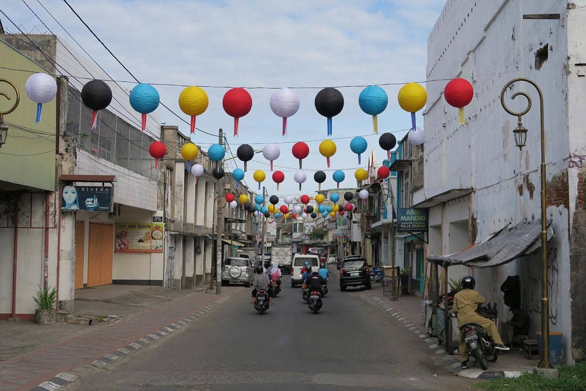 Deretan lampion warna-warni tergantung di atas jalan kota Mataram dengan kendaraan dan pengendara motor melintas di bawahnya.