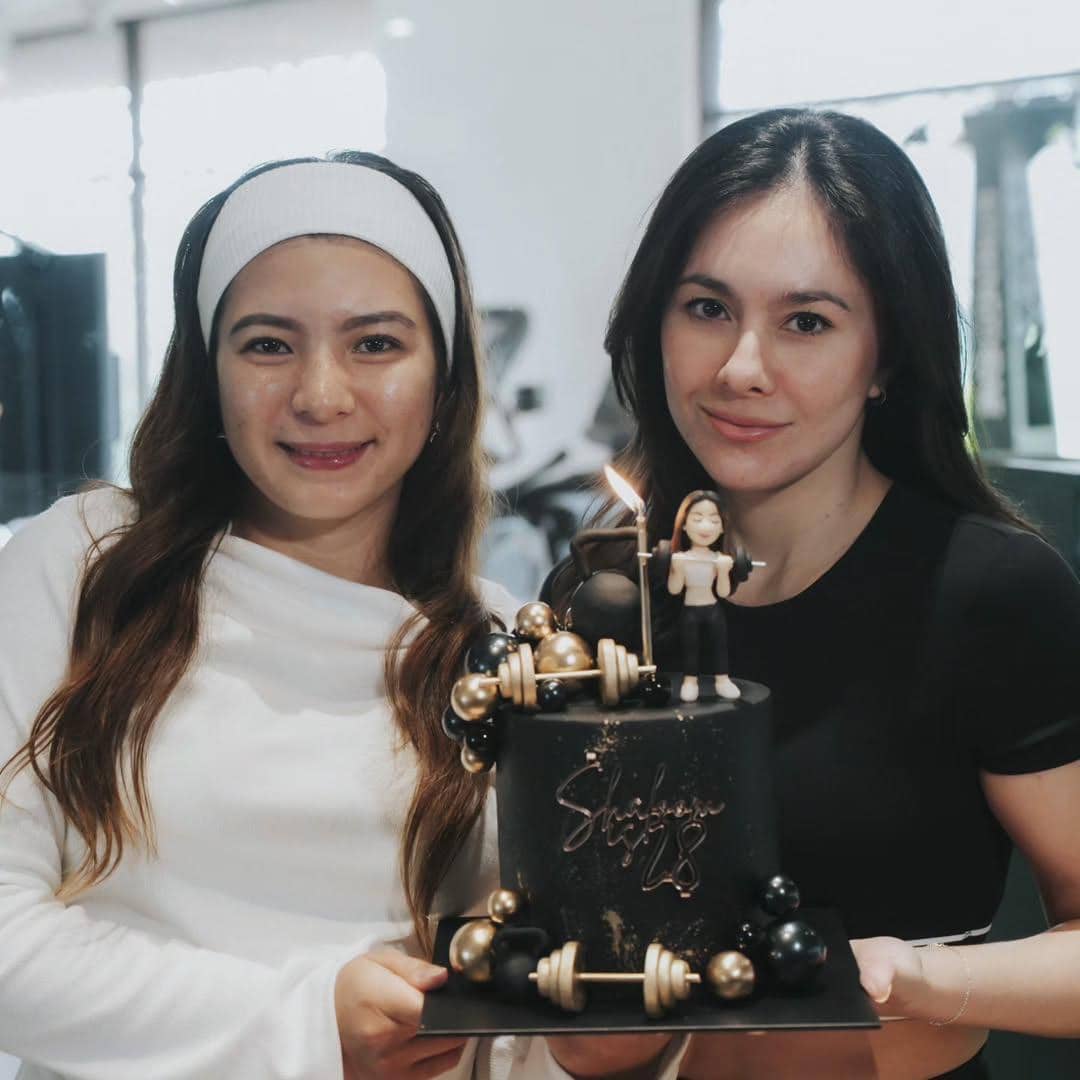 Dua perempuan tersenyum sambil memegang kue ulang tahun hitam dengan lilin menyala dan hiasan miniatur di atasnya.