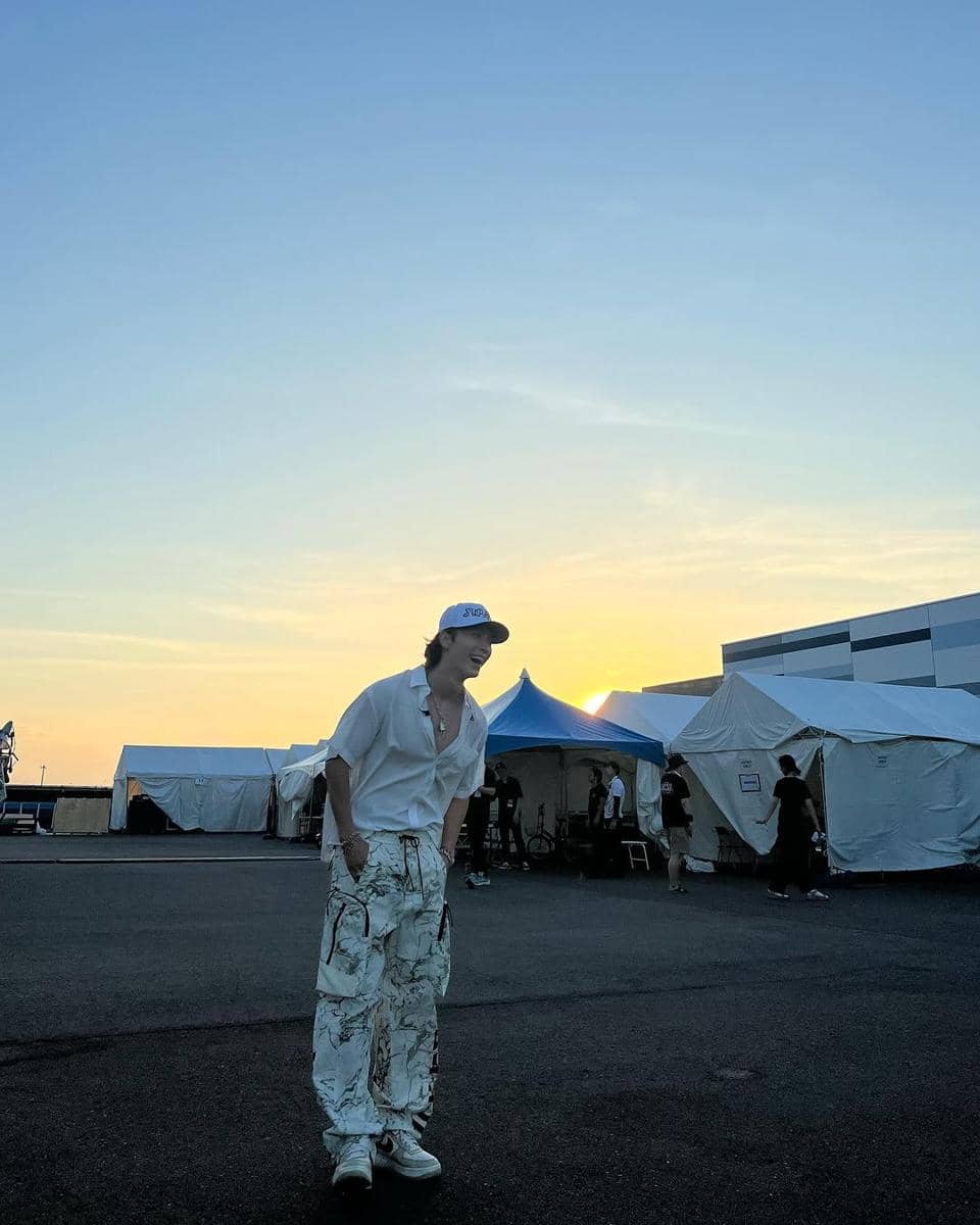 Seorang pria mengenakan kemeja putih longgar, celana kargo bermotif, dan topi putih berdiri di area tenda saat matahari terbenam.