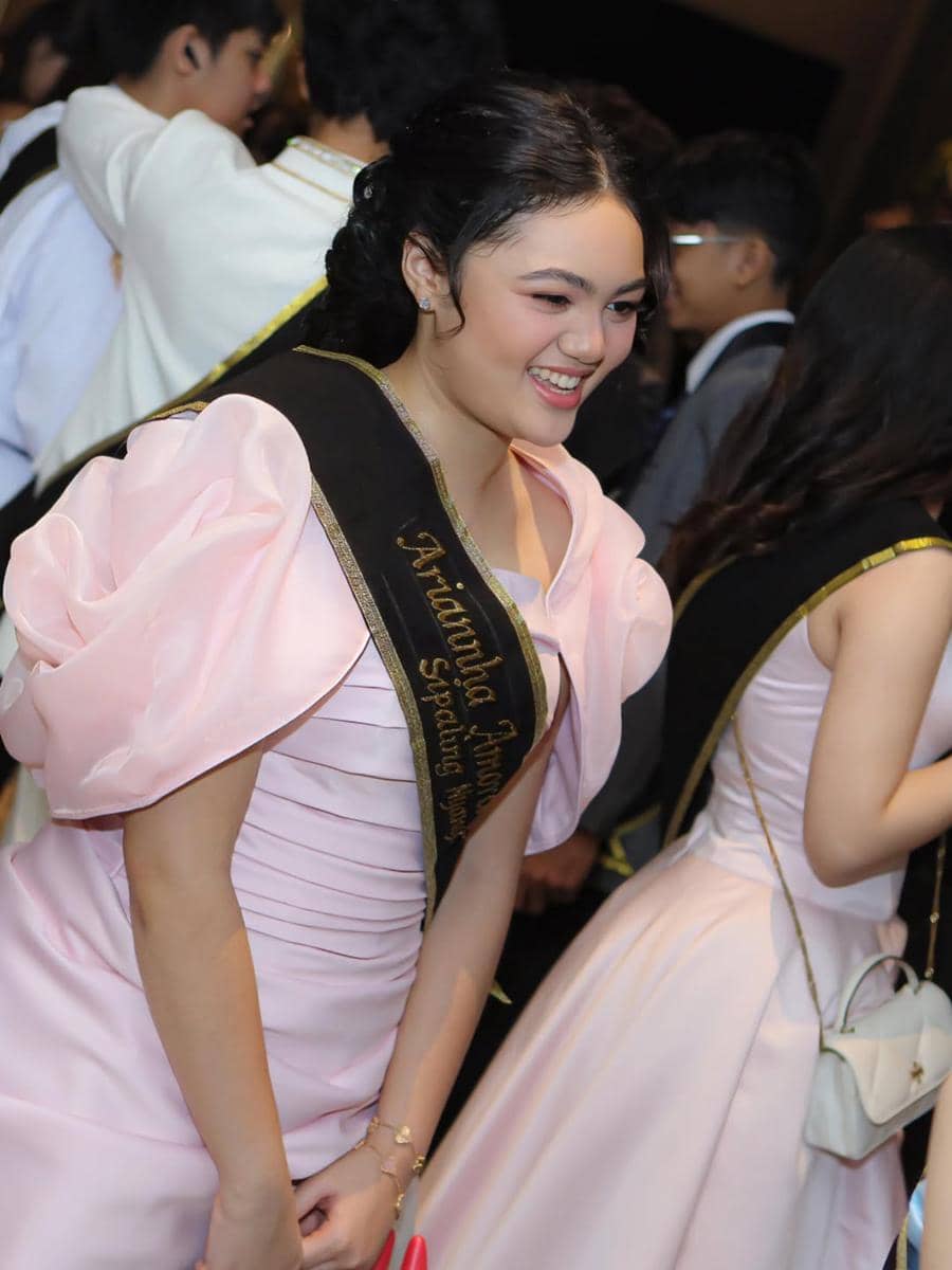 Seorang remaja mengenakan gaun berwarna merah muda dengan selempang hitam bertuliskan nama, tersenyum di acara prom night sekolah.