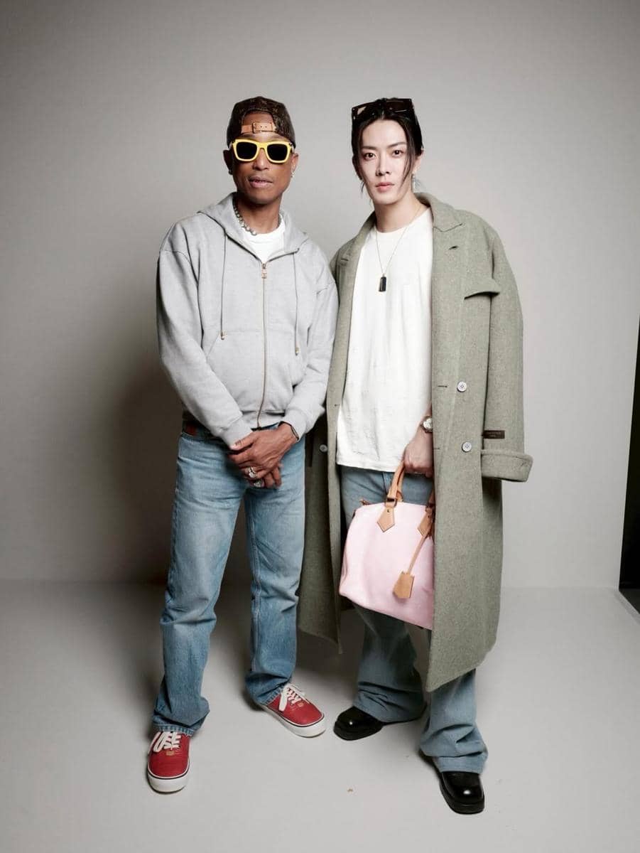 Dua pria berpose bersama di studio, satu mengenakan hoodie abu-abu dan kacamata kuning, satunya memakai mantel hijau dan membawa tas pink.