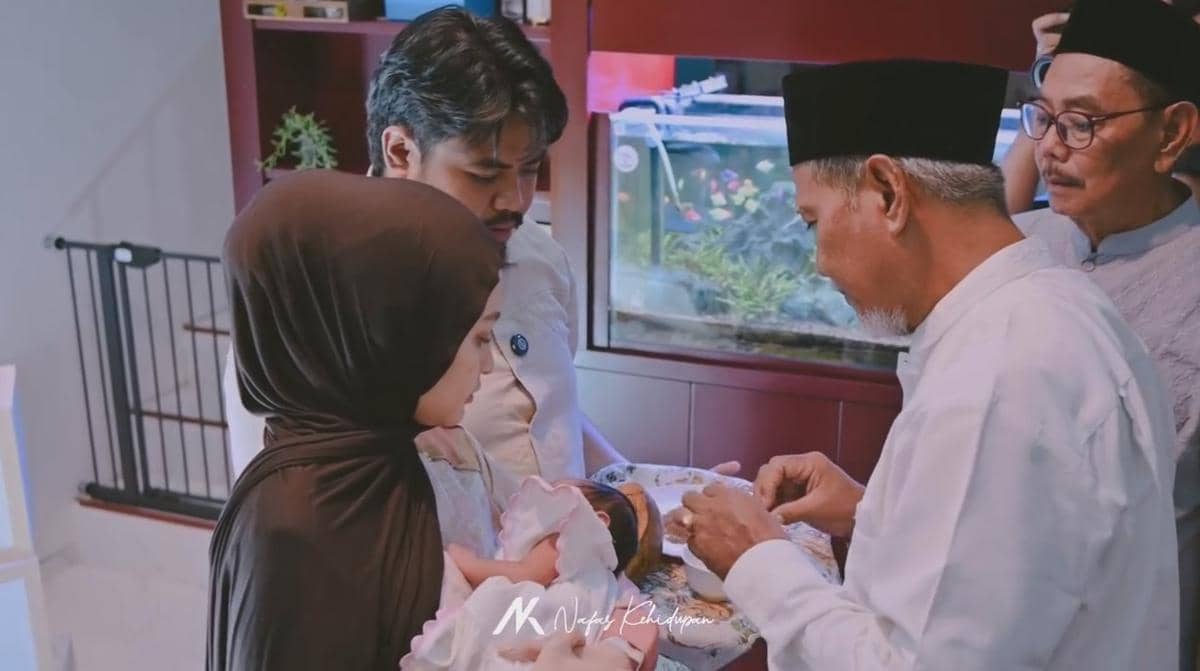 Seorang pria dan wanita menggendong bayi saat prosesi akikah, didampingi dua pria lanjut usia mengenakan peci di sebuah ruangan.