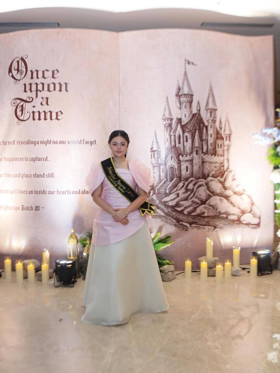 Seorang gadis mengenakan gaun elegan berwarna pink dan putih dengan selempang hitam berpose di depan latar bertema dongeng bertuliskan 'Once Upon a Time'.