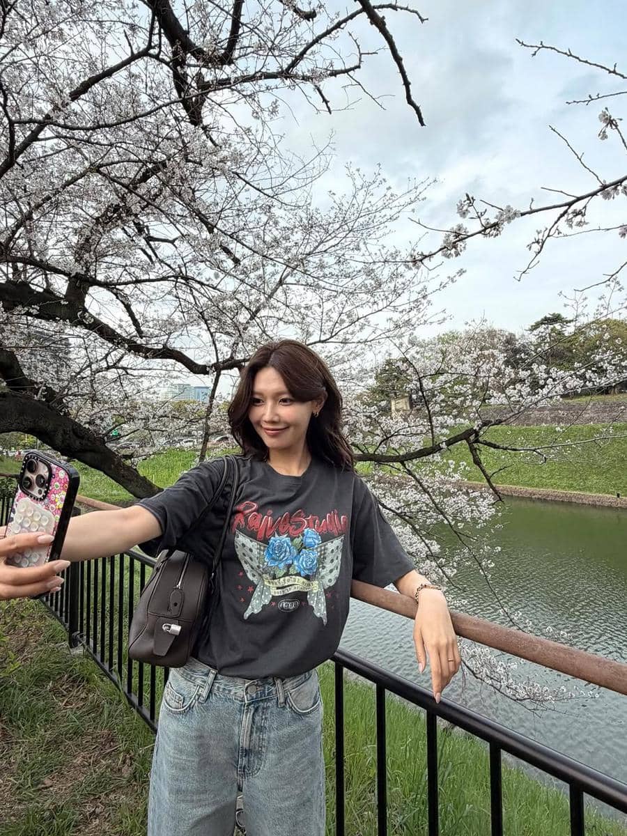 Seorang wanita berpose selfie di tepi sungai dengan latar pohon sakura bermekaran, mengenakan kaus abu-abu bergambar dan celana jeans longgar.